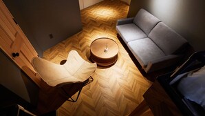 A compact room with a herringbone wooden floor, a grey sofa, a white chair, and a small round table with a lamp.