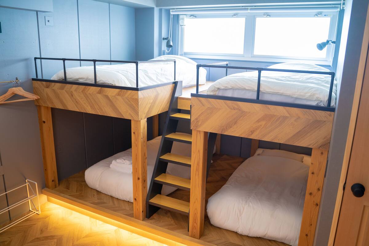 A bunk bed room with a wooden floor, a window with blinds, and a wall-mounted light fixture.