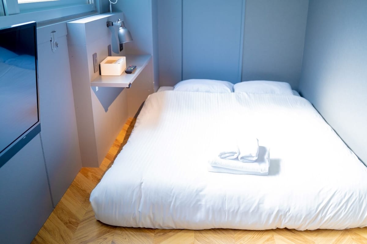 A small, neatly made bed with white linens and a folded towel, a bedside table with a lamp and a remote control, and a wooden floor.