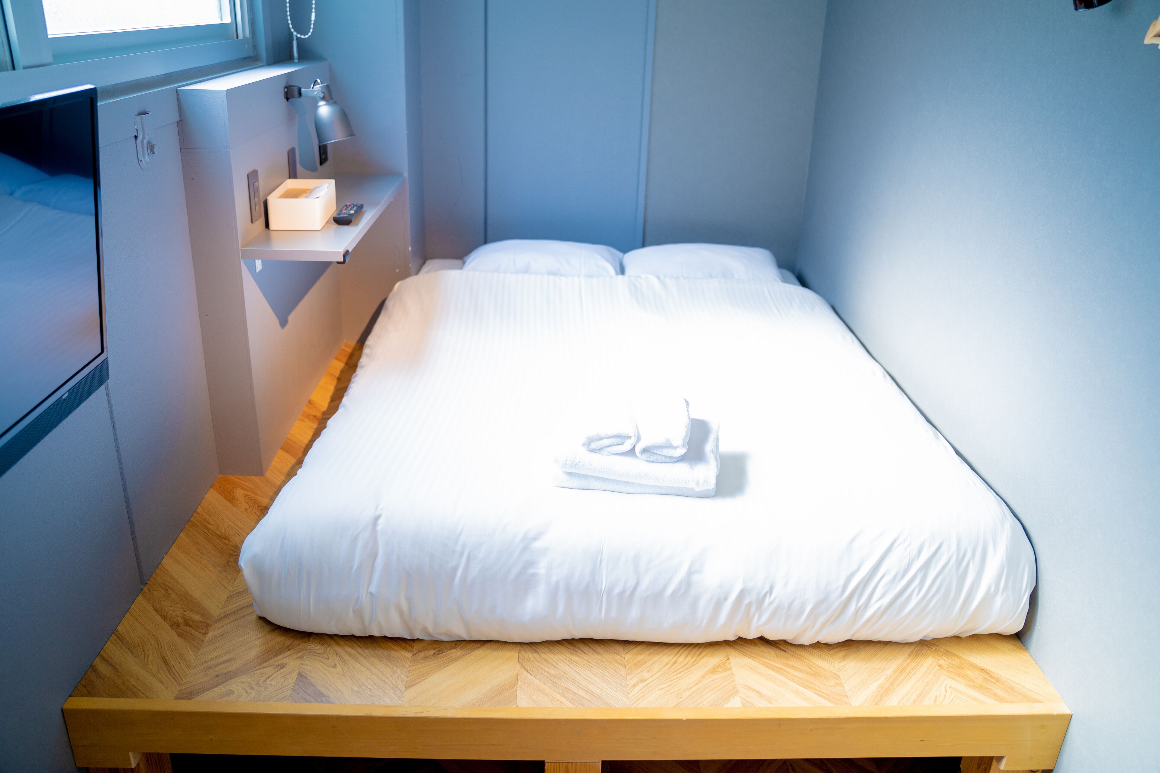 En pænt redt seng med hvide sengetæpper og et foldet håndklæde, et natbord med en lampe, og en lille hylde med en æske og en fjernbetjening.