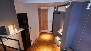 A compact room with a wooden herringbone floor, a bed, a desk with a chair, a wardrobe, and a small kitchenette area.
