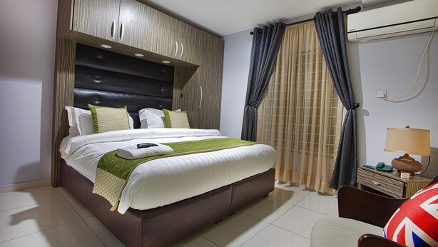 A neatly made bed with a headboard, a bedside table with a lamp, and a chair with a cushion. - H21 Apartments & Lounge (Lekki)