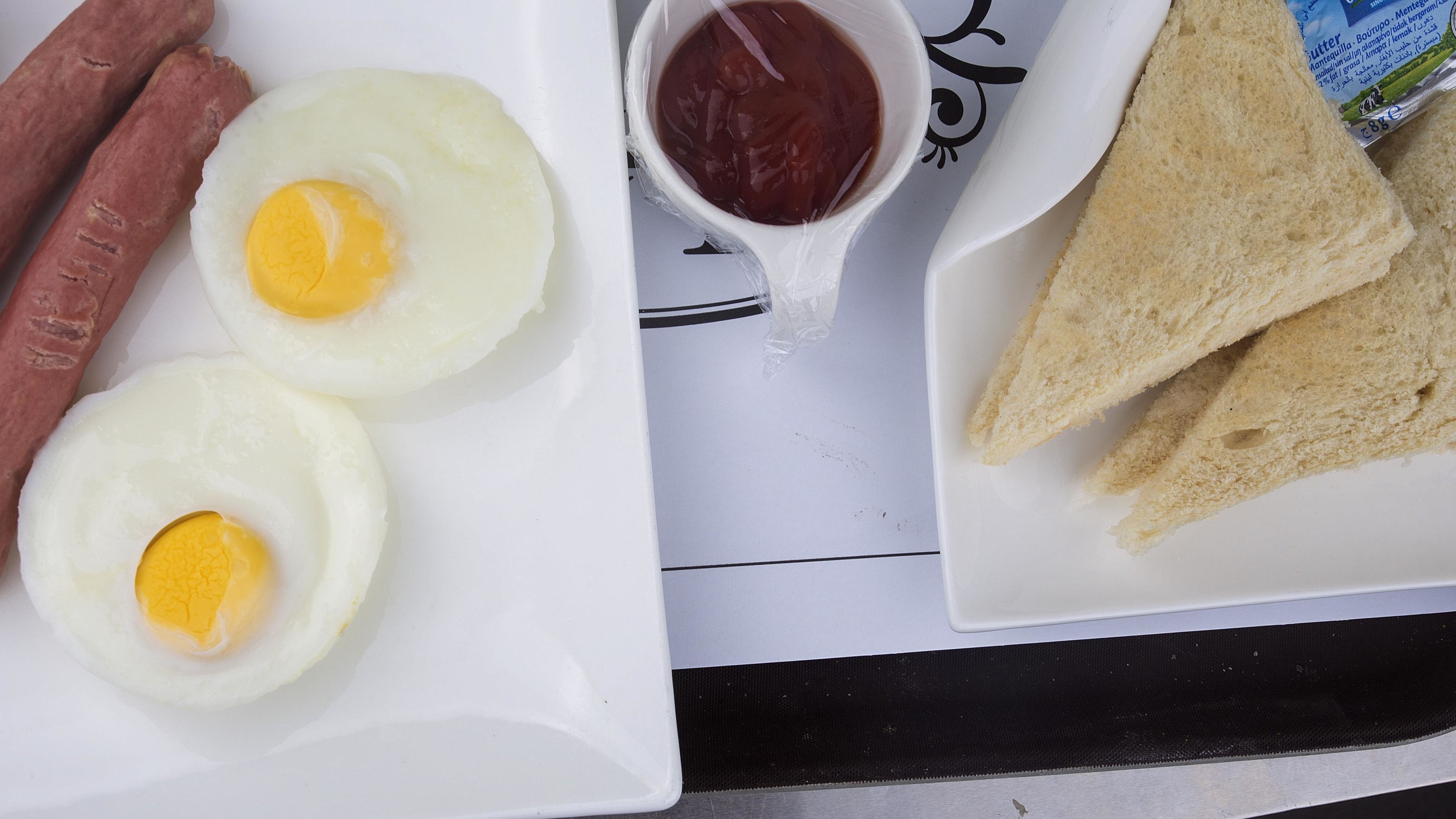 A plate with two fried eggs, two sausages, a small bowl of ketchup, and two slices of bread.