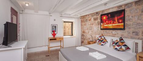 A bedroom with a bed, a desk, a TV, a window, and a stone wall.