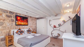 Une chambre avec un mur en pierre, un lit recouvert d’une couette grise, un téléviseur posé sur un meuble blanc et des livres sur une étagère incurvée.