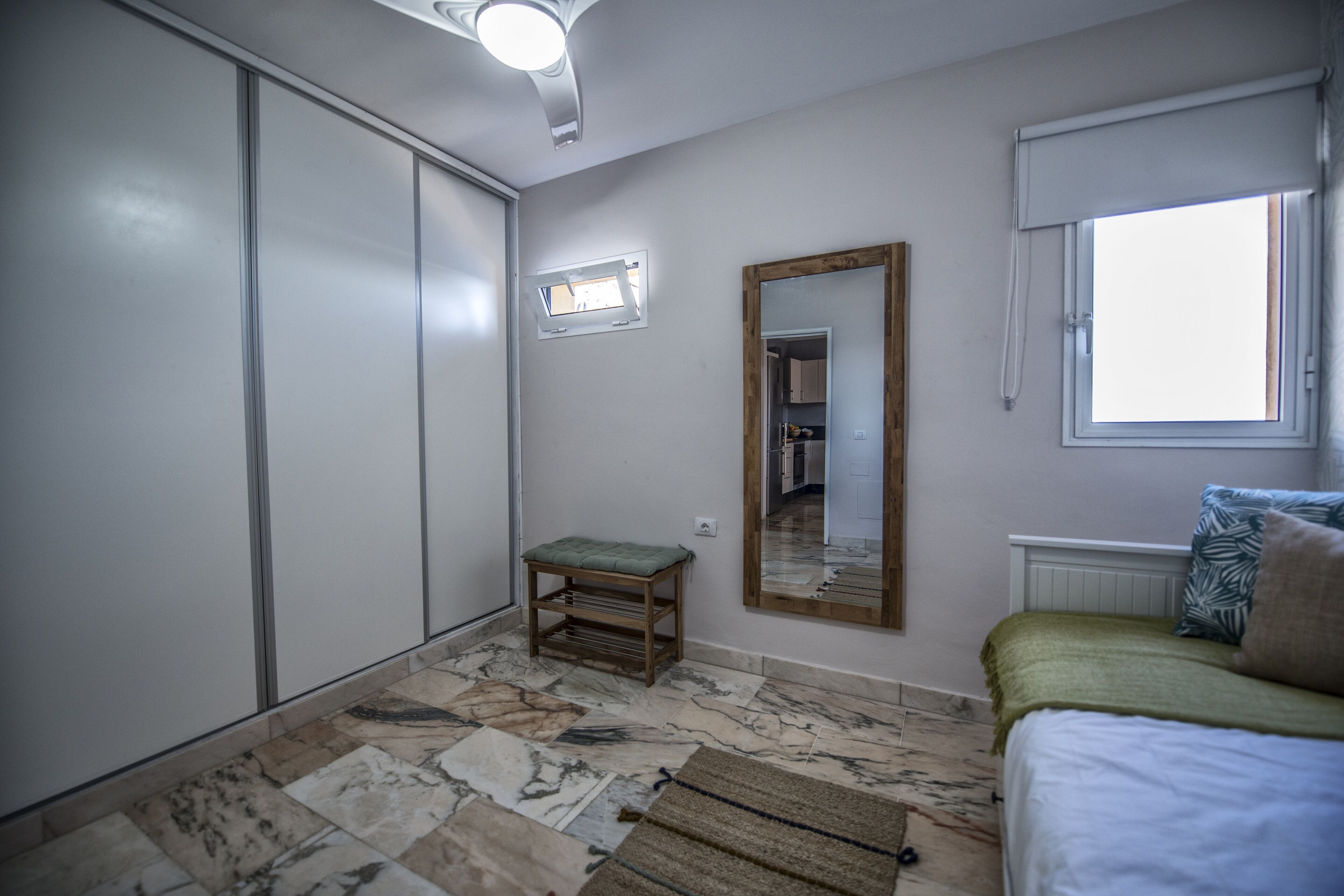 A bedroom with a bed, a window, a mirror, a wardrobe, and a small stool.