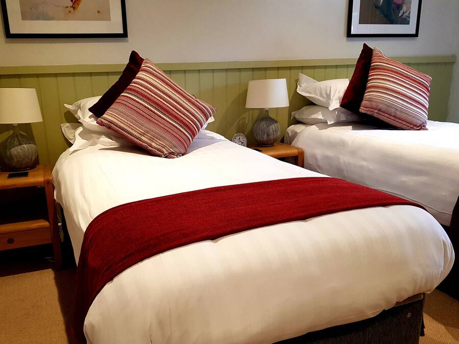 Une chambre d’hôtel avec deux lits, chacun agrémenté de draps blancs et de plaids rouges. On y trouve des tables de chevet avec des lampes et une horloge. La chambre est dotée d’une tête de lit verte et d’un tableau encadré au mur.