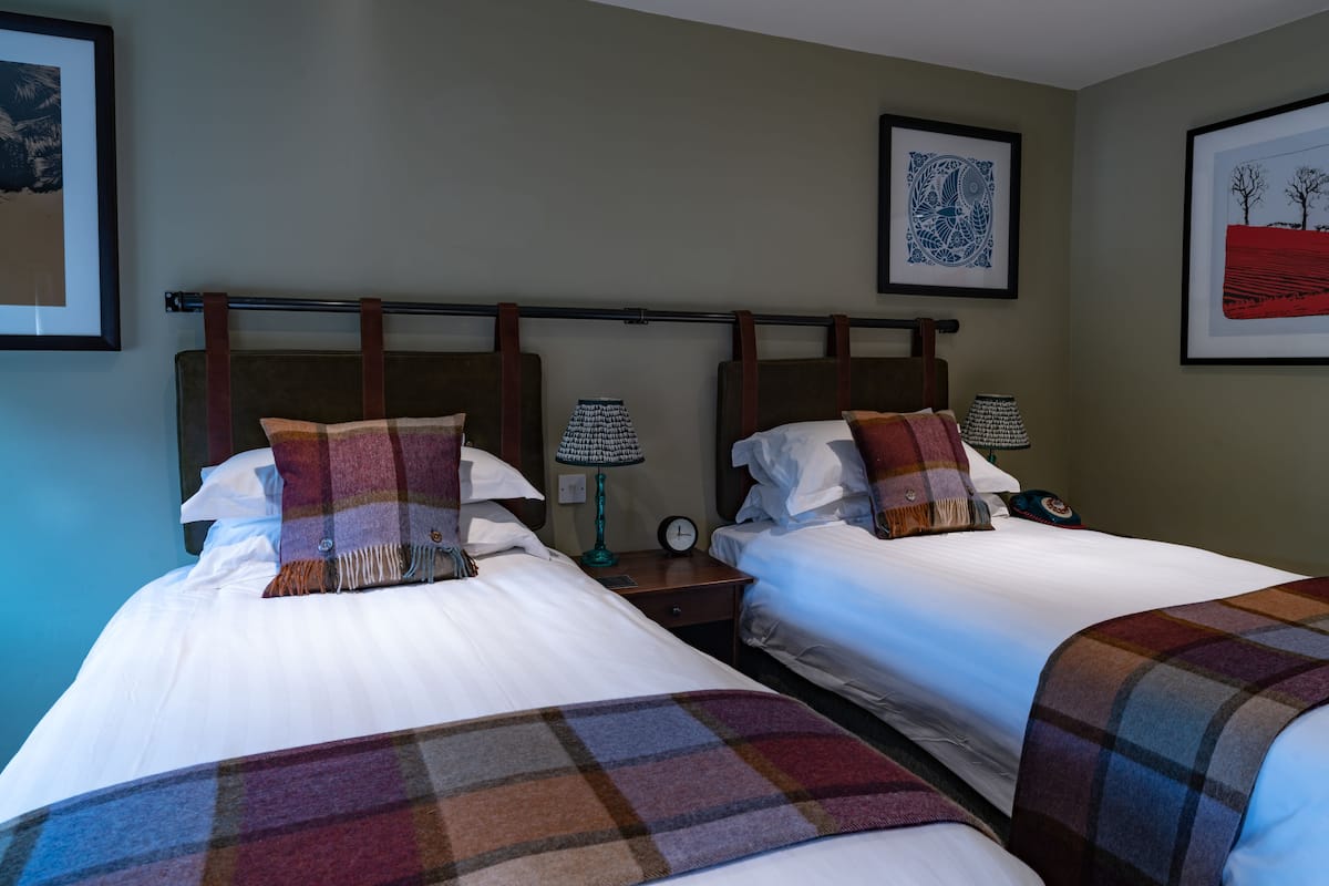 Habitación de hotel con dos camas, cada una con manta y almohadas a cuadros, una mesita de noche con lámpara y cuadros en las paredes.