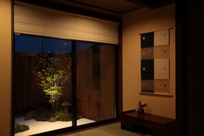 Una habitación con puerta de vidrio corredera que da a un jardín con un árbol bonsái, una pequeña mesa con un florero y un suelo de esteras de tatami.