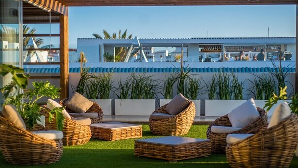 A rooftop terrace with wicker furniture, artificial grass, and a view of a bar and buildings in the background. - Sunset Apartment (Corralejo Fuerteventura) (Corralejo)