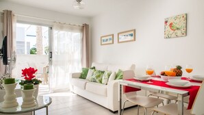 A modern living room with a white sofa, a dining table with red tablecloth, and a glass coffee table. There are two framed pictures on the wall, a ceiling light, and a window with curtains. - Sunset Apartment (Corralejo Fuerteventura) (Corralejo)