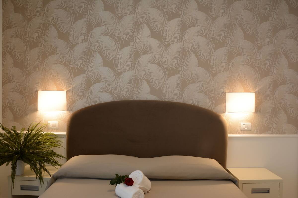 A neatly made bed with a headboard, two bedside lamps, a potted plant, and a rolled towel with a rose on a nightstand.