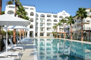Un área de piscina con sillones de descanso y sombrillas, un edificio con balcones y palmeras.