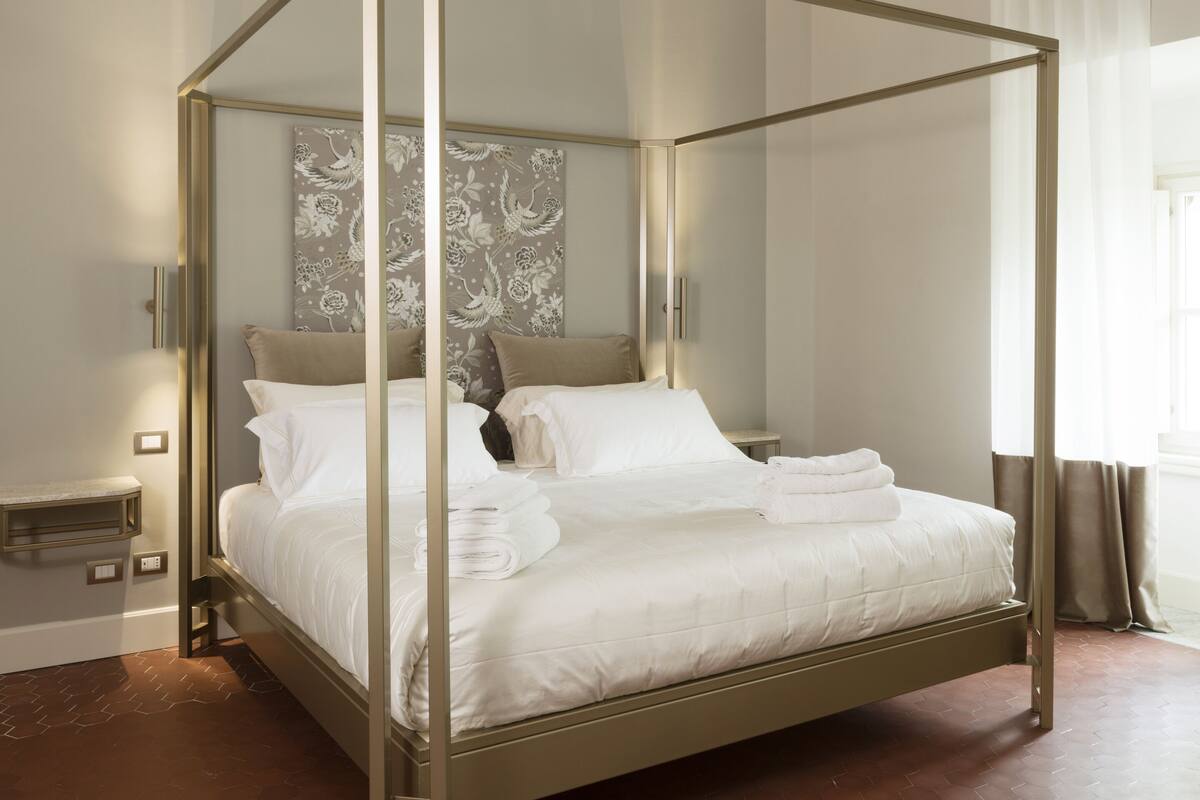 A four-poster bed with a white bedspread and pillows, a nightstand with a lamp, and a wall-mounted artwork.