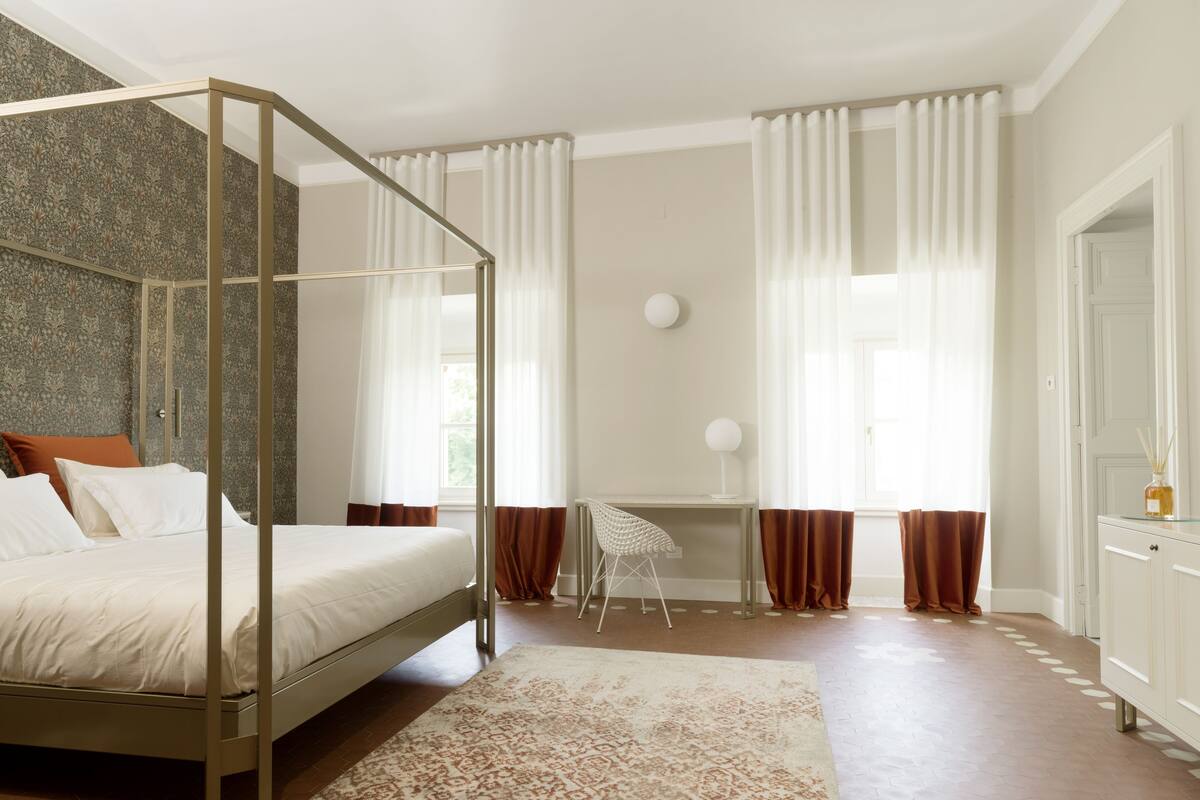 A four-poster bed with a headboard, a desk with a chair, a radiator, and a window with curtains.