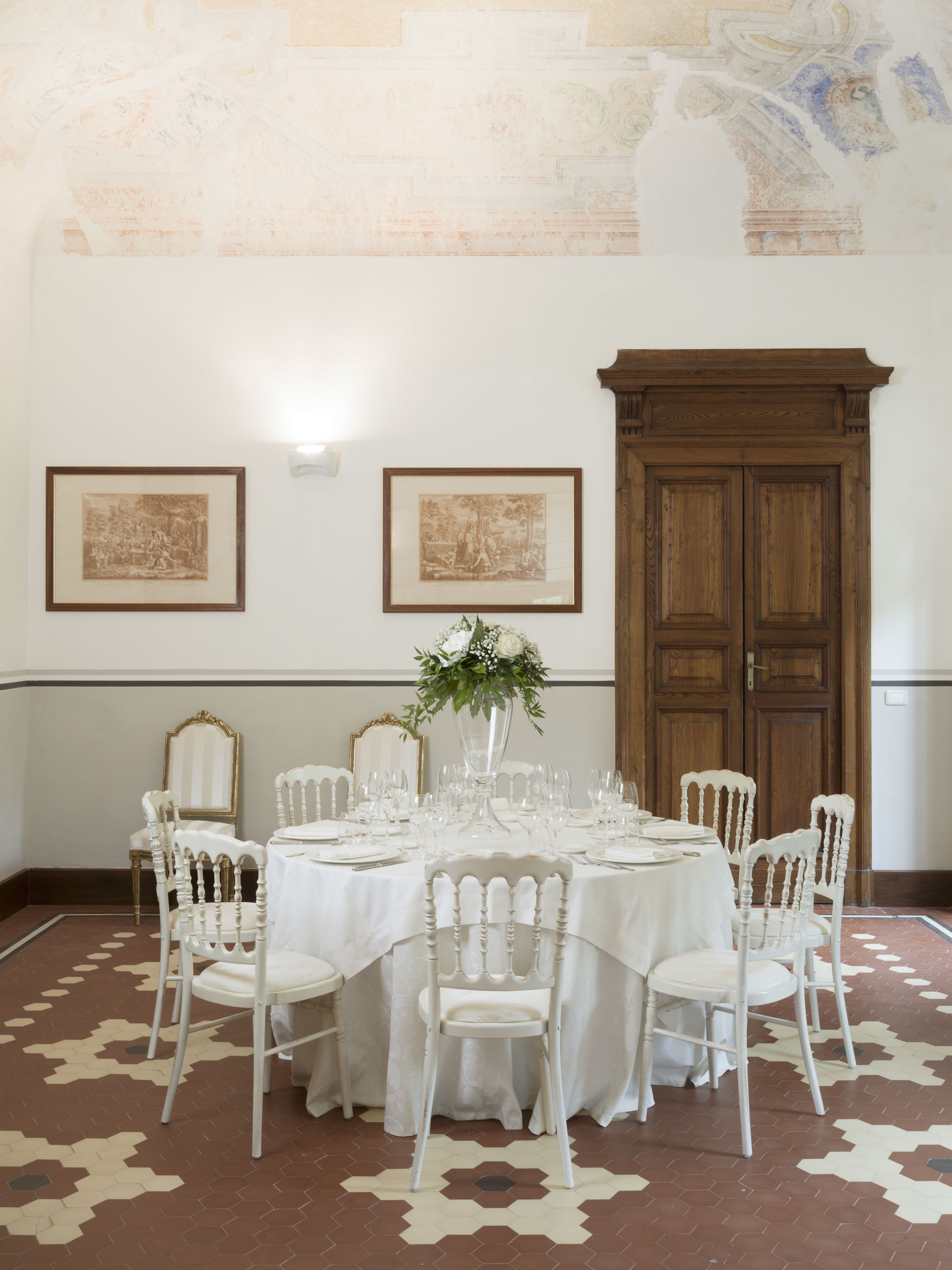 A dining room with a round table set for six, surrounded by white chairs, a vase with flowers, and two framed pictures on the wall.
