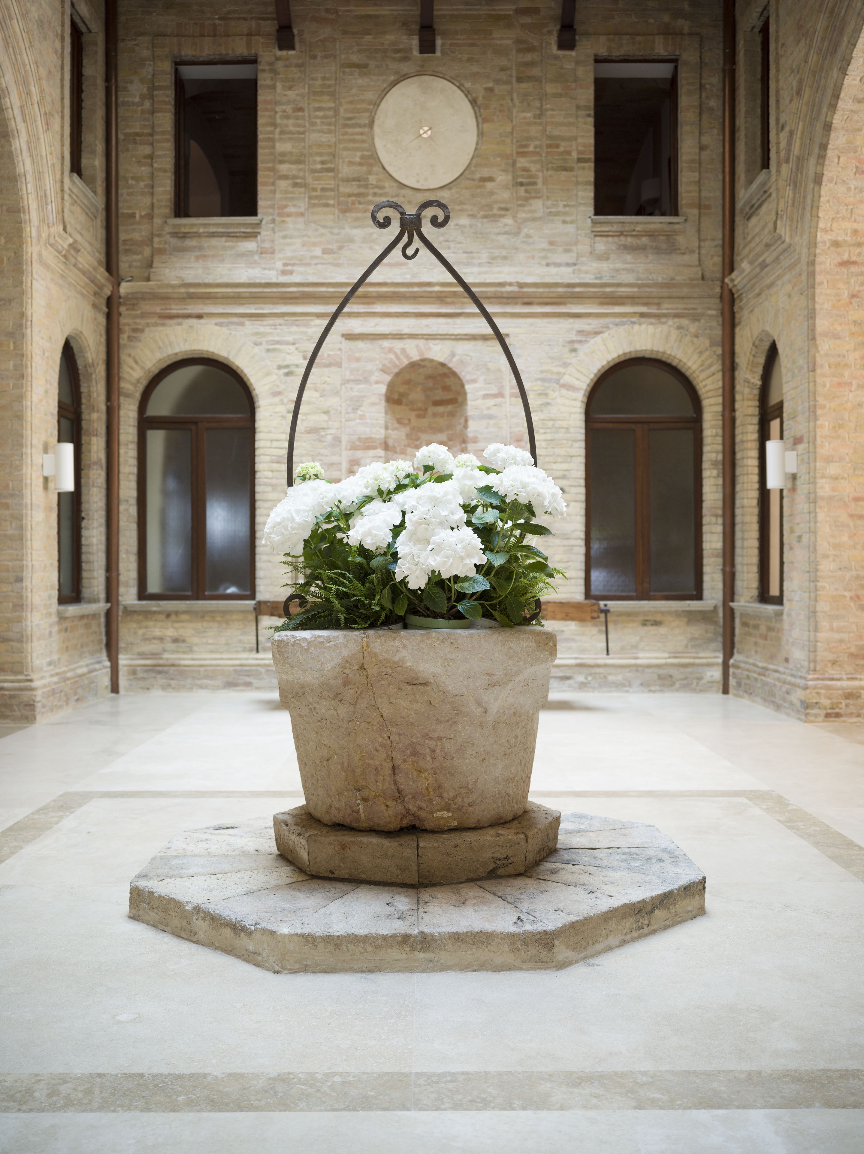 A large stone planter with white flowers in a spacious, arched room with brick walls and a clock on the wall.