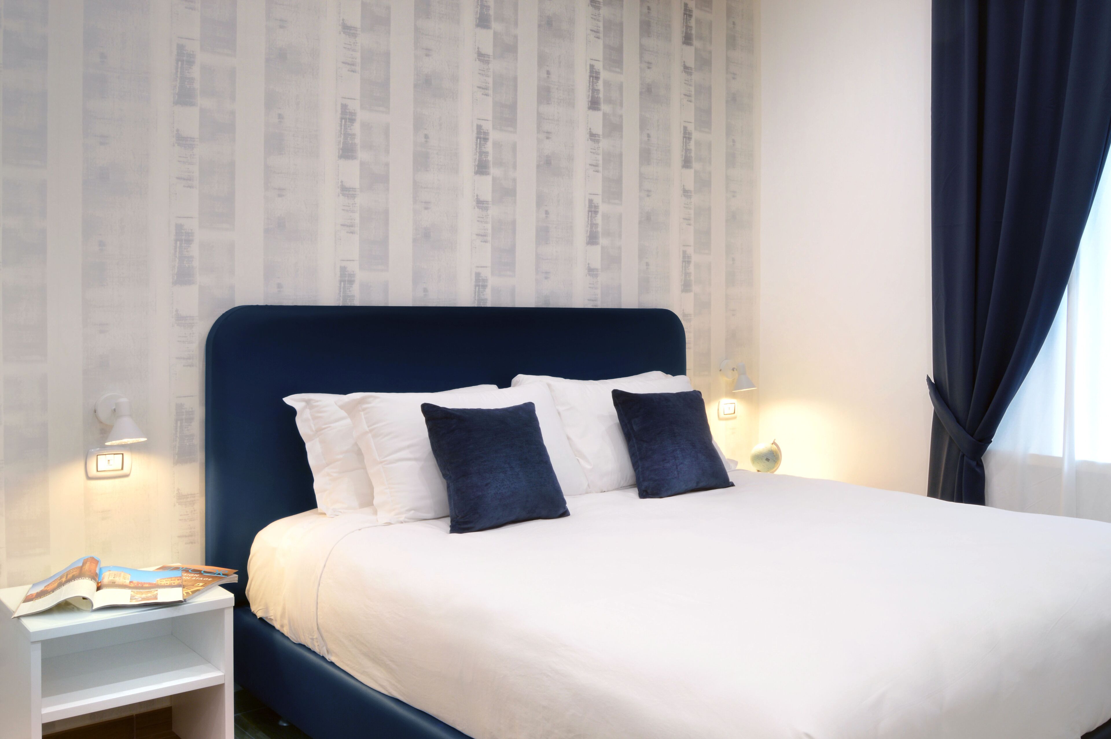 A neatly made bed with white and dark blue pillows, a white nightstand with a magazine, and a wall with vertical stripes.
