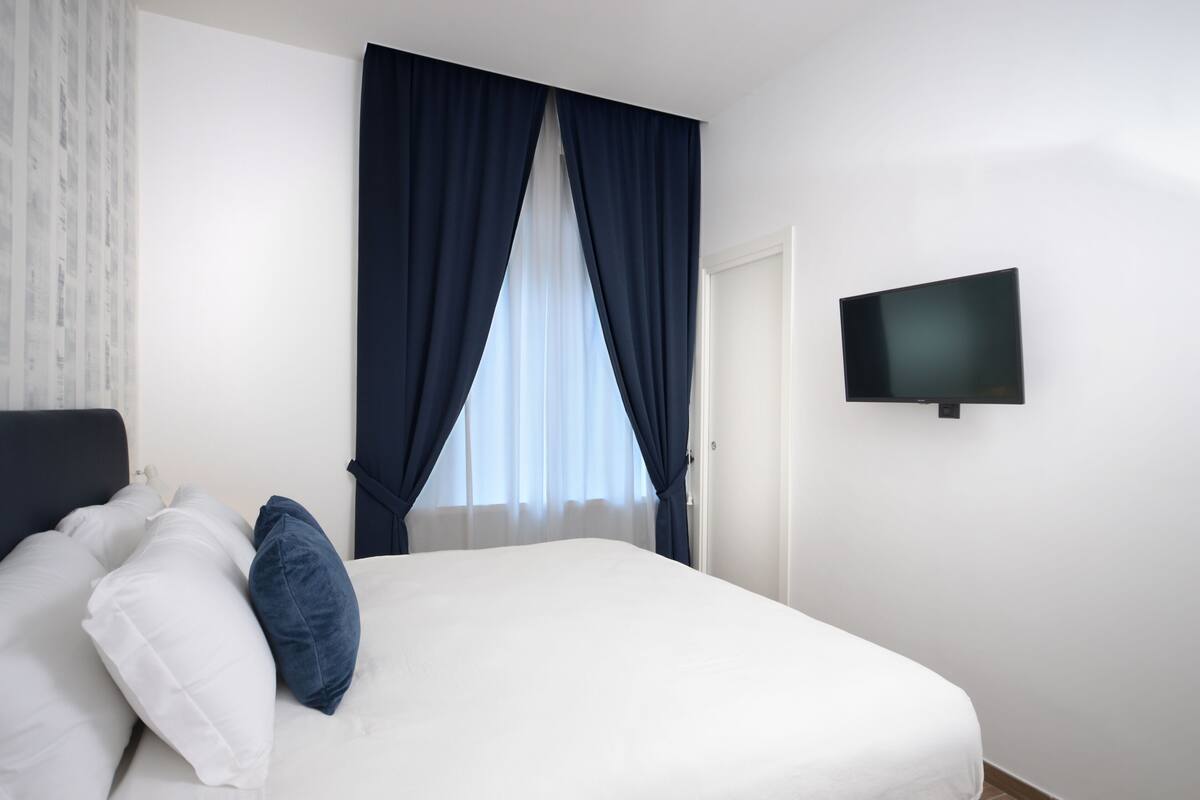A bedroom with a bed, white bedding, a TV mounted on the wall, and a window with blue curtains.