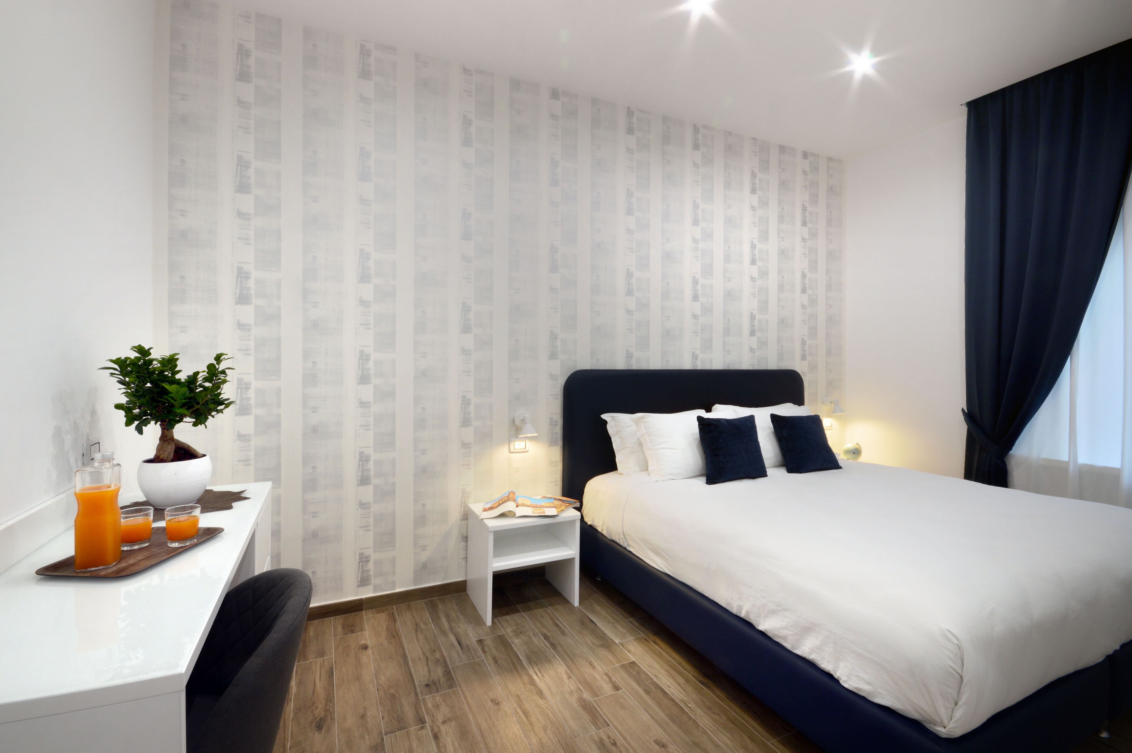 A hotel room with a bed, a desk with a plant, a small nightstand, and a window with curtains.