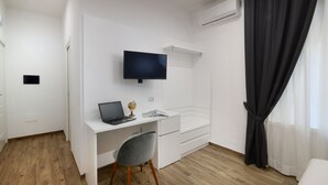 Une chambre d’hôtel moderne avec un bureau blanc, un téléviseur à écran plat, un ordinateur portable et une chaise grise.