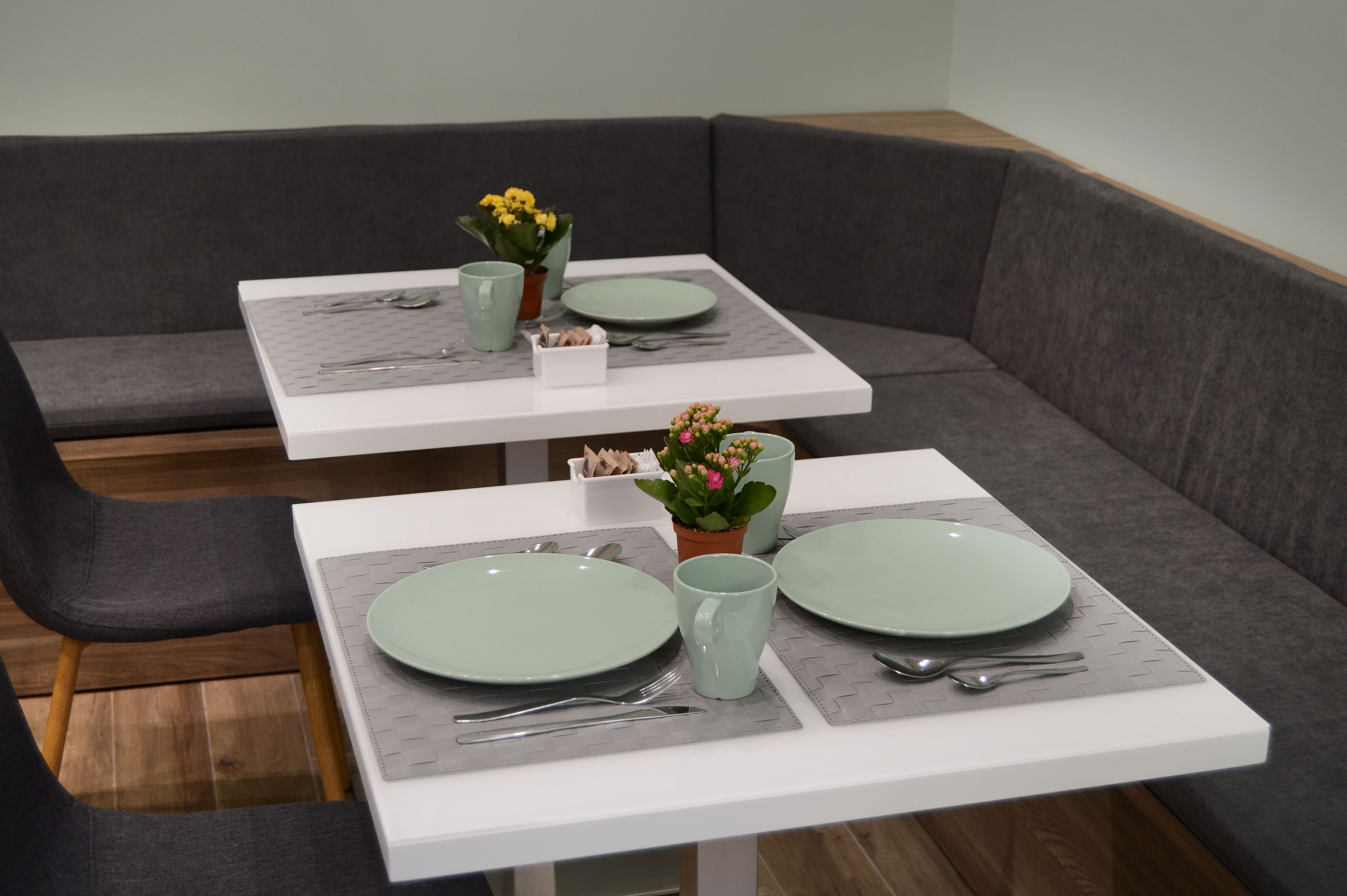 Un área de comedor con dos mesas cuadradas, cada una con un plato verde, una taza y cubiertos. Hay plantas en macetas sobre las mesas.