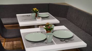 Un área de comedor con dos mesas cuadradas, cada una con un plato verde, una taza y cubiertos. Hay plantas en macetas sobre las mesas.