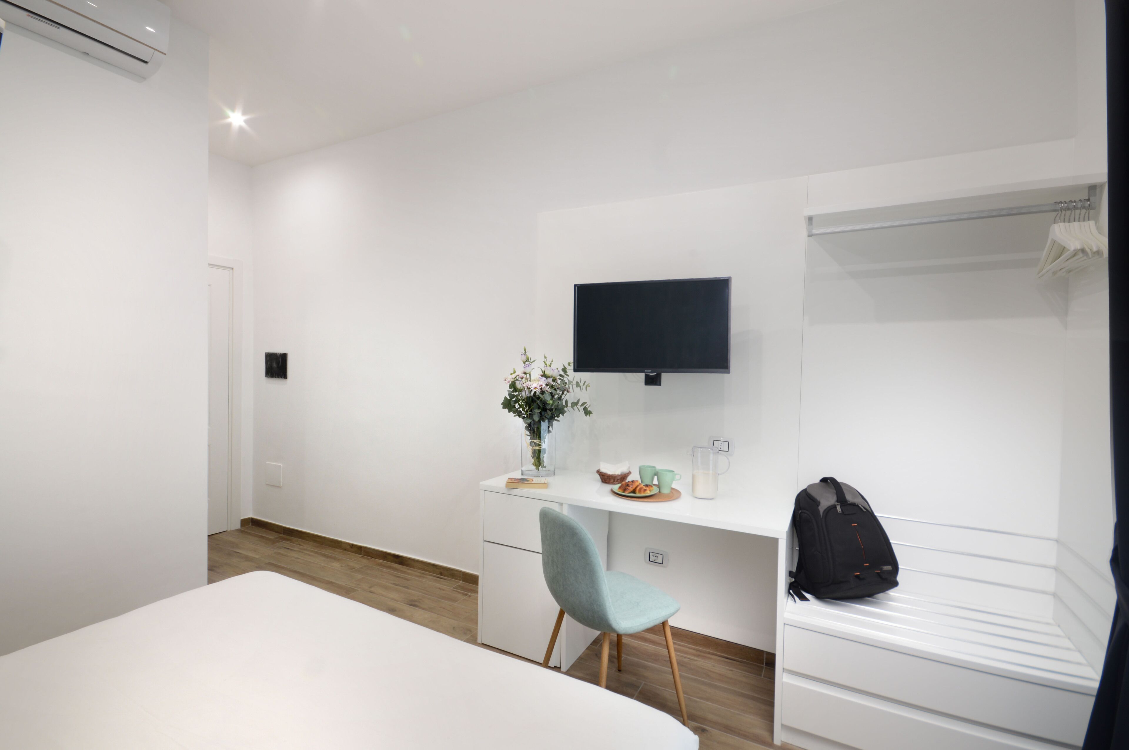 Una camera d'albergo moderna con un letto, una scrivania con televisore a schermo piatto, una sedia, un piccolo tavolo con un vaso di fiori e un armadio.