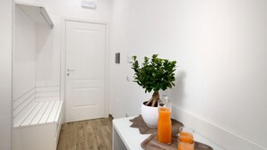 Un couloir avec une porte blanche, une plante en pot, un comptoir blanc sur lequel se trouvent un verre de jus d’orange et un autre verre de jus d’orange, le tout posé sur un plateau en bois.