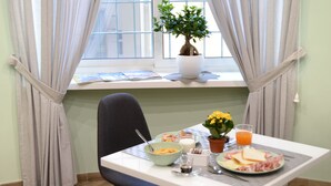 Une table dressée pour le petit-déjeuner, avec une chaise, des aliments pour le petit-déjeuner, une plante en pot et une fenêtre avec des rideaux.
