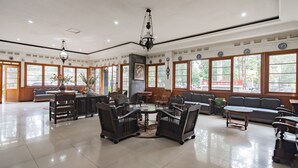 A spacious hotel lobby with a central round table surrounded by chairs, large windows with wooden frames, and a chandelier.