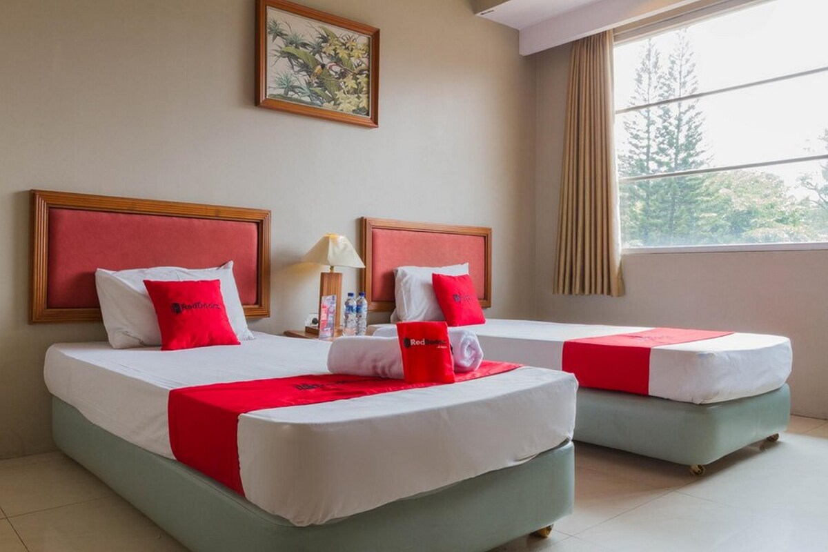 Ein Hotelzimmer mit zwei Betten, jeweils mit rotem Kopfteil, weißer Bettwäsche und einer roten Decke. An der Wand hängt ein Gemälde, es gibt ein Fenster mit Vorhängen und einen kleinen Tisch mit Lampe und Flaschen.