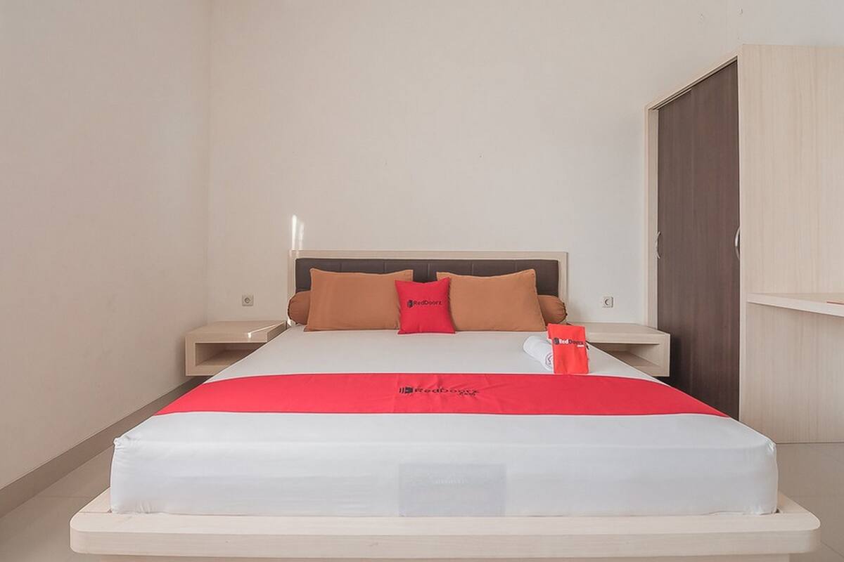 Ein ordentlich bezogenes Bett mit einer roten und weißen Tagesdecke, zwei Kissen und einem Kissen mit einem Logo. Neben dem Bett steht ein Nachttisch mit einer Lampe und einem kleinen Tisch.
