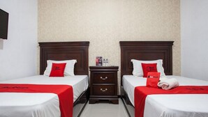 A hotel room with two beds, each with a red bedspread and pillows, a wooden nightstand with a bottle of water, and a wall with a subtle pattern.