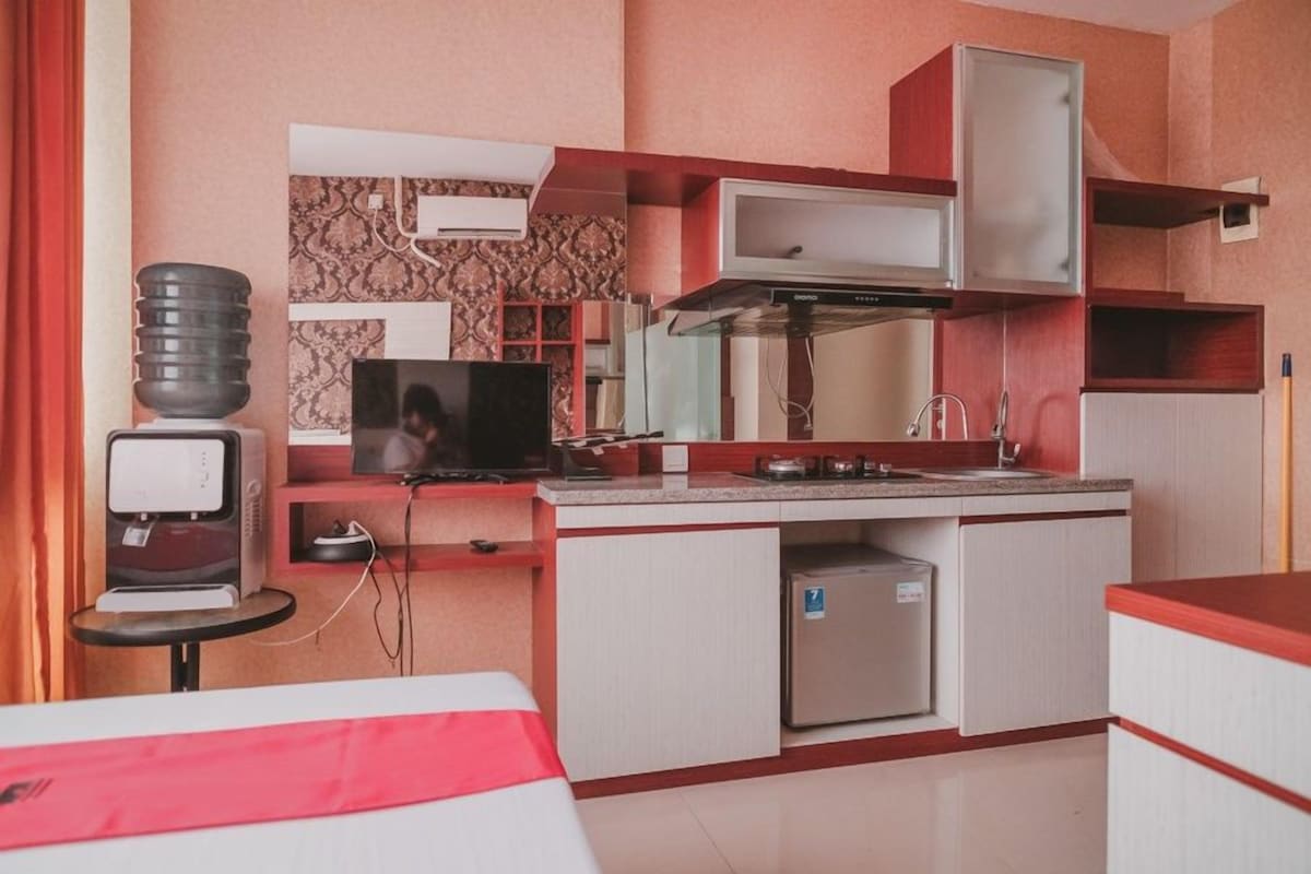 Habitación de hotel pequeña con kitchenette, televisión, refrigerador y cama con detalles en rojo.