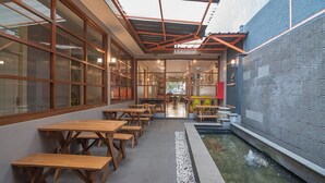 Un comedor interior con mesas y bancos de madera, una pequeña fuente de agua y vistas al exterior a través de grandes ventanales.