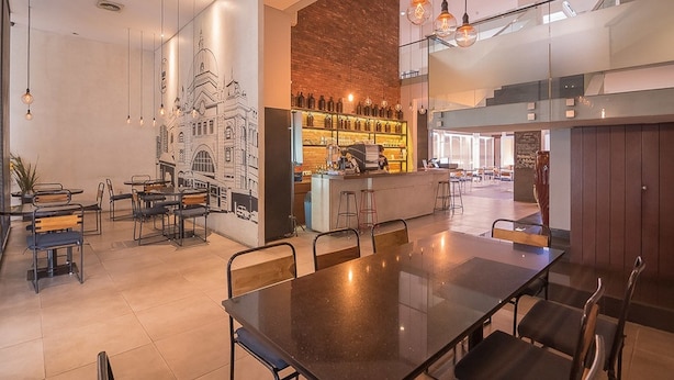 A modern dining area with a long table and chairs, a bar with shelves of bottles, and a mural of a building on the wall. - RedDoorz Plus @ Cipaganti Street 3 (Bandung)