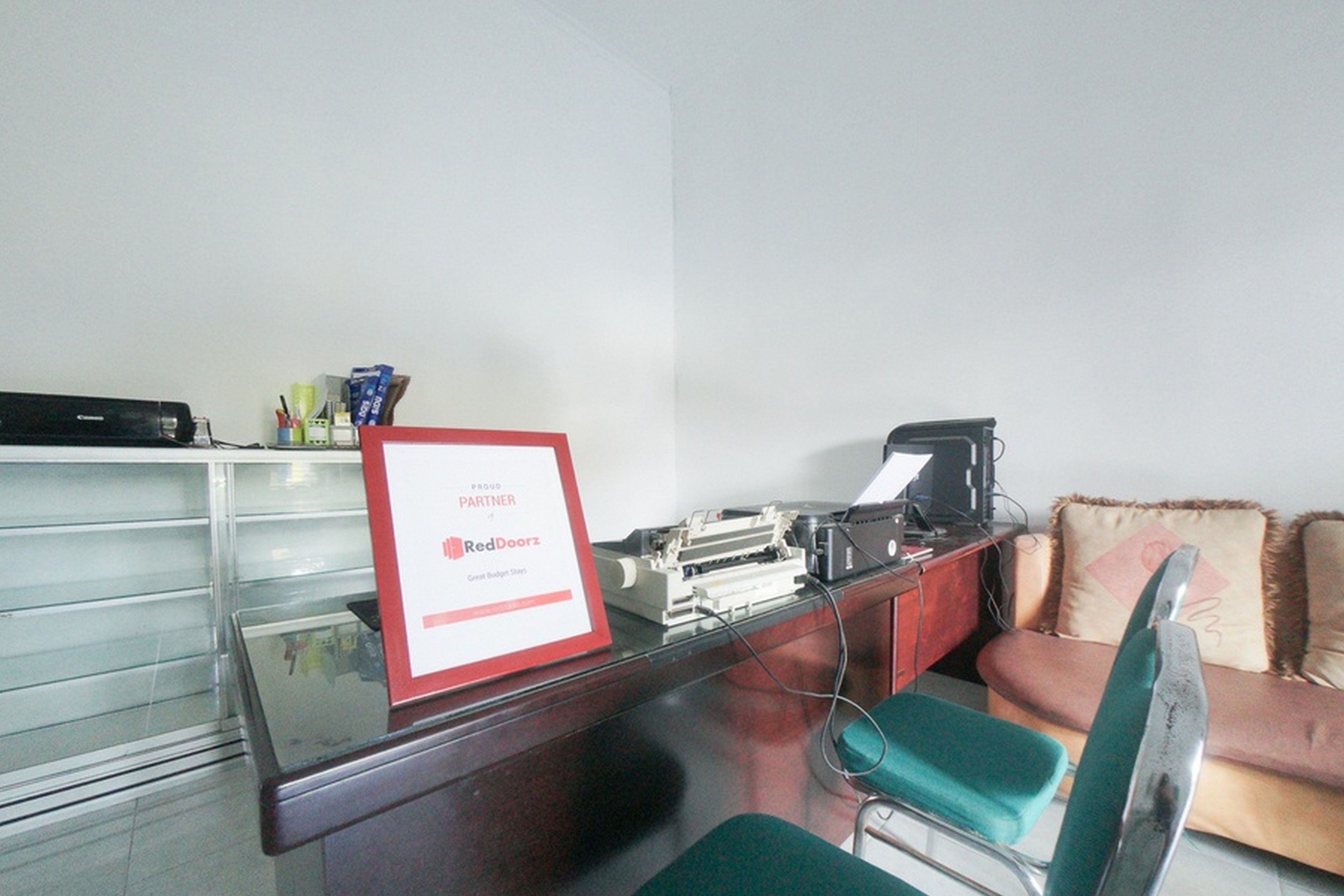 A room with a desk, a computer, a printer, a sofa, and a sign indicating a partnership with RedDoorz.