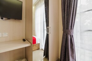 A hotel room with a TV mounted on the wall, a bedside table, a bed with a red blanket, and a window with curtains. - Residences by RedDoorz @ Guntur Raya Setiabudi (Jakarta)