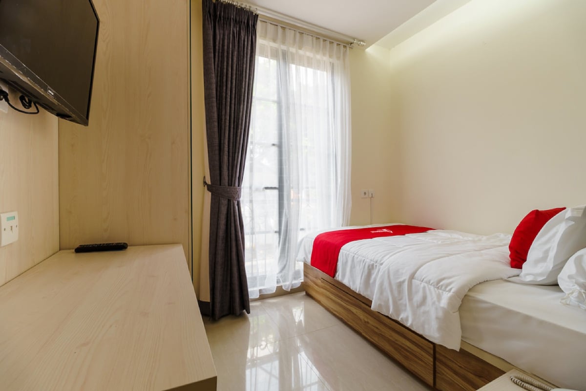 A bedroom with a bed, a desk, a TV mounted on the wall, and a window with curtains.
