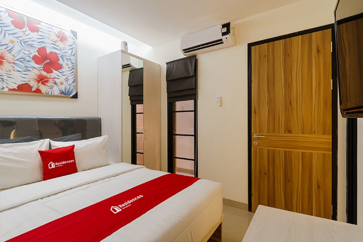 A hotel room with a bed, a red and white bedspread, a red pillow, a wooden door, and a wall-mounted air conditioner.