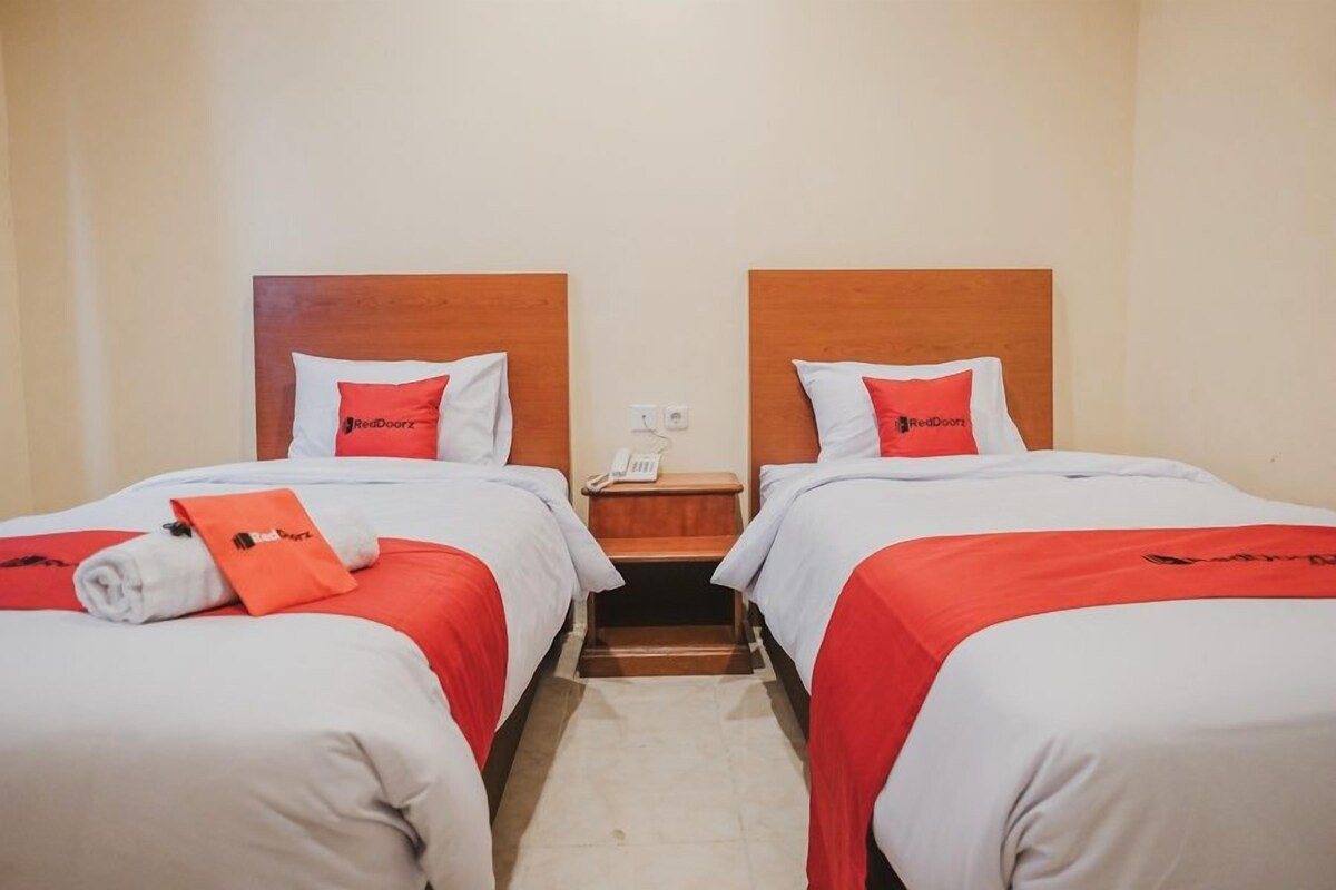 Ein Hotelzimmer mit zwei Betten, jeweils mit einem hölzernen Kopfteil, weißer Bettwäsche und roten Akzenten. Zwischen den Betten befindet sich ein Nachttisch mit einem Telefon und einer kleinen Ablagefläche.