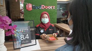 A reception desk with a framed customer review award, a RedDoorz logo, and a woman in a red headscarf holding a tray. - RedDoorz Plus near Plaza Blok M (Jakarta)