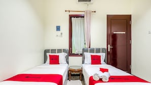 Une chambre d’hôtel avec deux lits simples, chacun doté d’une tête de lit rouge et de draps blancs. Un petit tabouret en bois se trouve entre les lits, et il y a une fenêtre avec des rideaux blancs.
