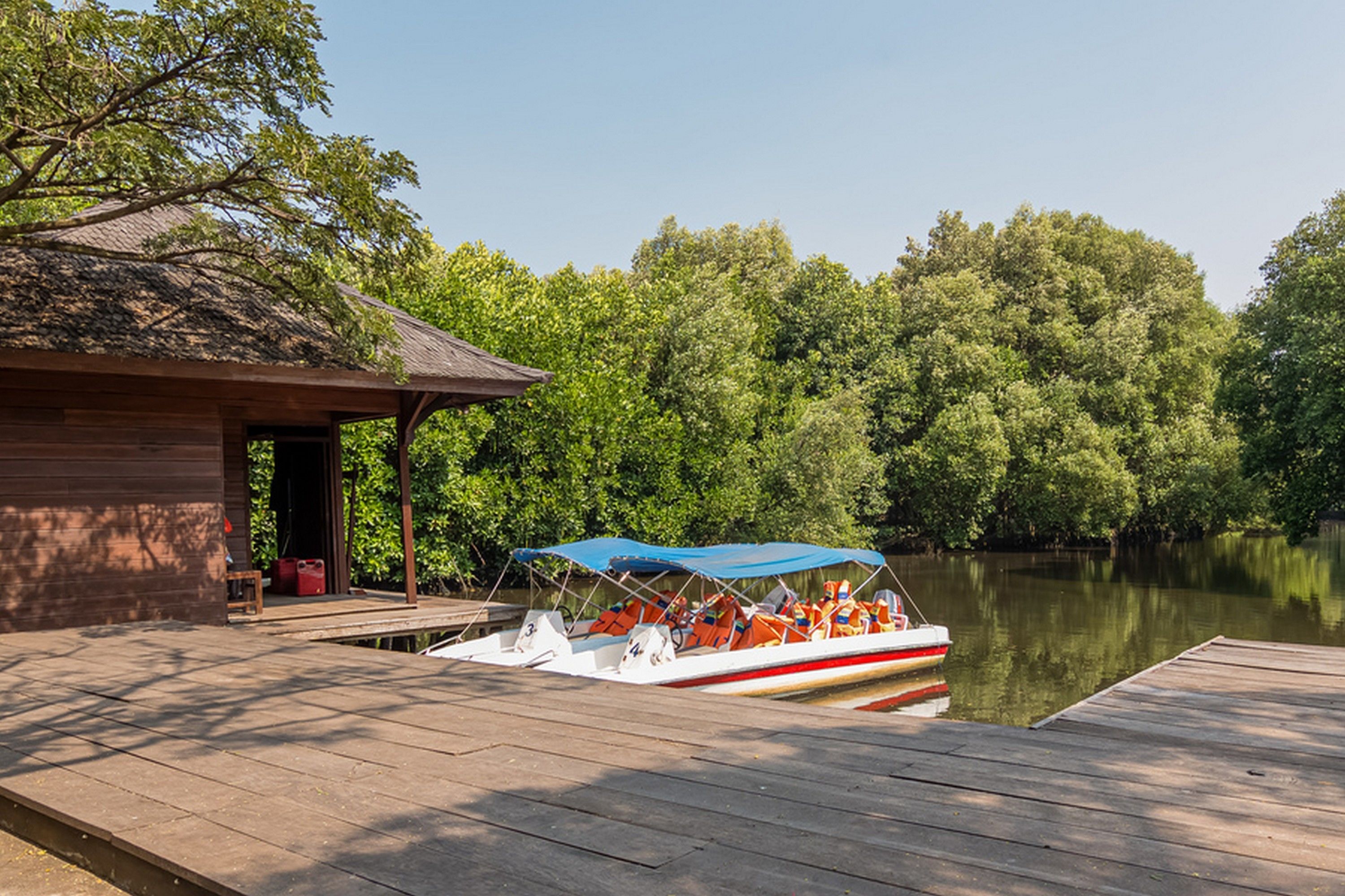 Une jetée en bois avec un bateau recouvert d’une bâche bleue, un bâtiment en bois doté d’un toit en pente, et, au fond, de nombreux arbres verdoyants.