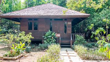 A wooden house with a tiled roof, surrounded by lush greenery and a garden path leading to the entrance.