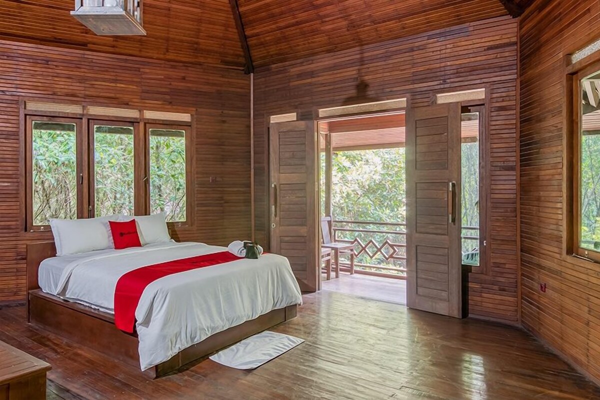 A wooden cabin bedroom with a bed, a red throw blanket, and a balcony with a wooden railing.