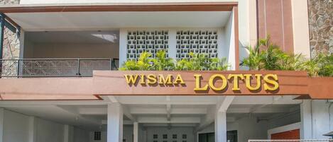 Un bâtiment sur lequel on peut lire l’inscription « WISMA LOTUS », et une zone de stationnement couverte où se trouvent une voiture et des motos.