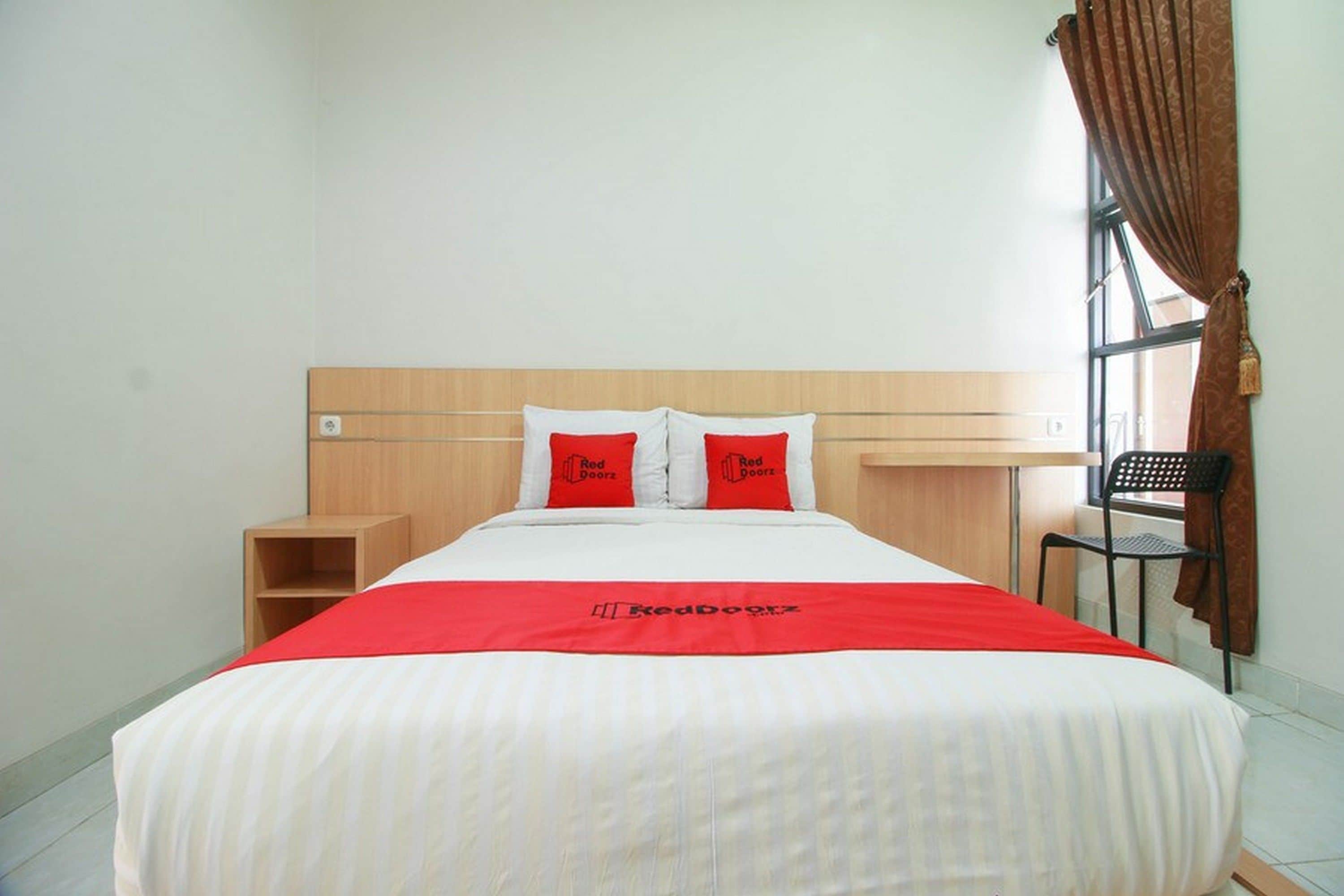 A hotel room with a bed, a wooden headboard, a red and white bedspread, two red pillows, a nightstand, a chair, and a window with curtains.