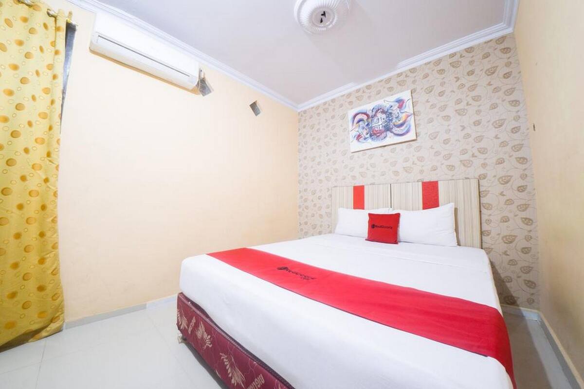 Habitación de hotel con cama, colcha roja y blanca, cortina amarilla, pared con estampado floral y ventilador de techo.